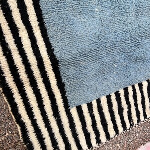 May include: A close-up of a blue and white striped rug. The rug has a light blue center and a border of black and white vertical stripes. The rug's texture appears soft and plush.