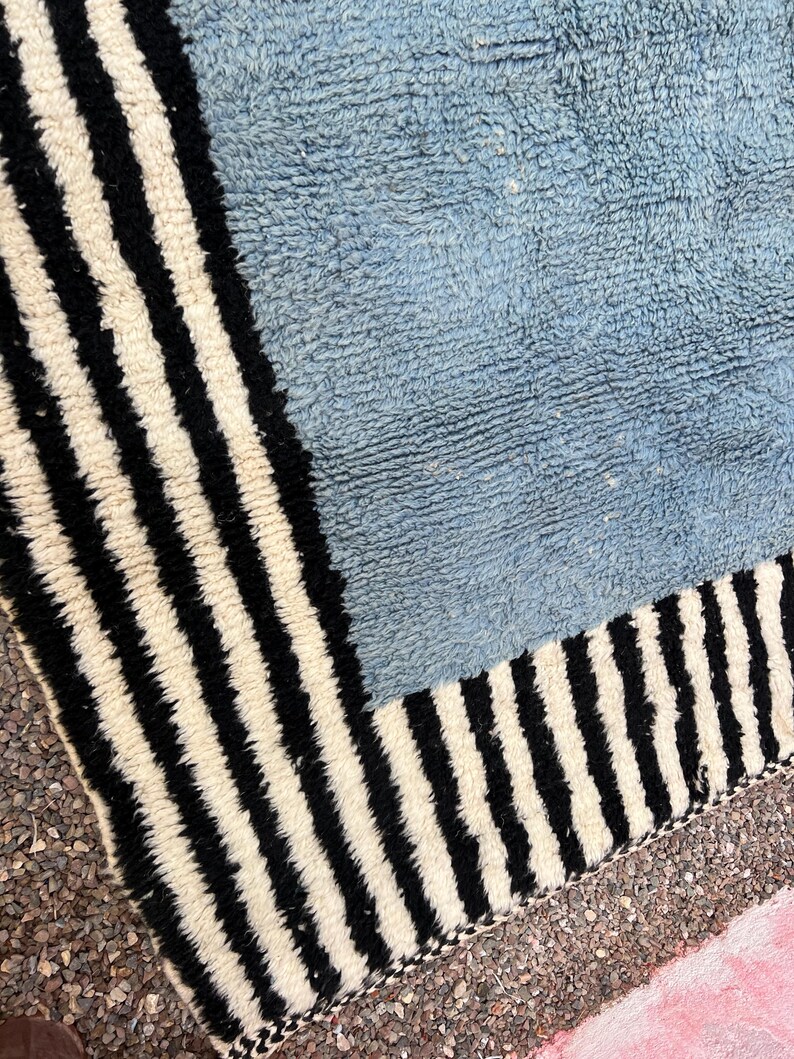 May include: A close-up of a blue and white striped rug. The rug has a light blue center and a border of black and white vertical stripes. The rug's texture appears soft and plush.
