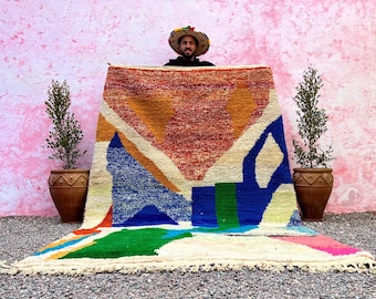 May include: A colorful Moroccan rug with geometric shapes in shades of blue, green, orange, and red. The rug is laid out on a gravel surface in front of a pink wall.
