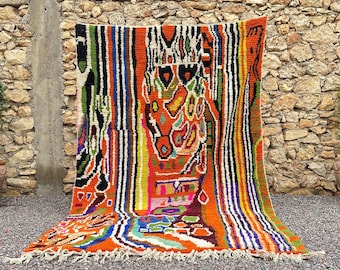 May include: A vibrant, hand-woven rug with a bold, abstract design. The rug features a mix of colors, including orange, green, blue, and white, arranged in vertical stripes and geometric shapes. The rug has a fringed edge and is displayed against a stone wall.