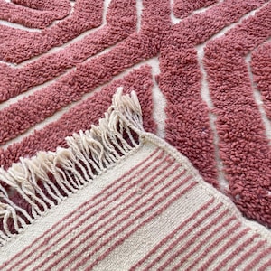 May include: A pink and white woven rug with a geometric pattern and fringe. The rug has a soft, textured surface.