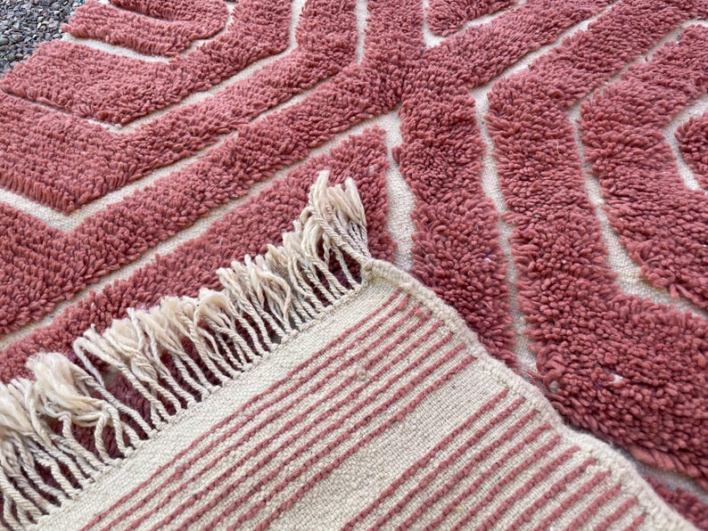 May include: A pink and white woven rug with a geometric pattern and fringe. The rug has a soft, textured surface.