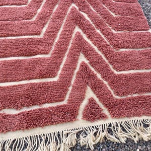 May include: A pink and white geometric patterned rug with a fringe border. The rug has a chevron pattern with white lines on a pink background.