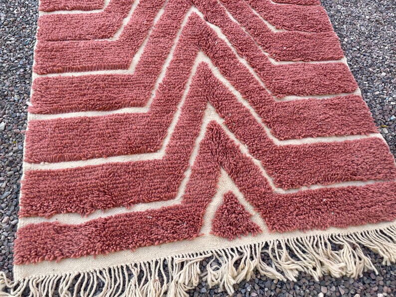 May include: A pink and white geometric patterned rug with a fringe border. The rug has a chevron pattern with white lines on a pink background.