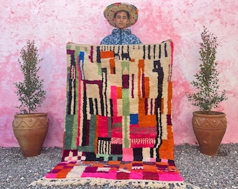 May include: A colorful Moroccan rug with geometric patterns in shades of pink, orange, green, blue, and black. The rug has a fringe border and is being held up by a person wearing a straw hat.