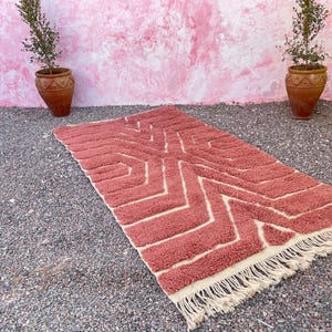 May include: A pink and cream patterned rug with a geometric design. The rug has a fringe border and is laid out on a gravel surface.