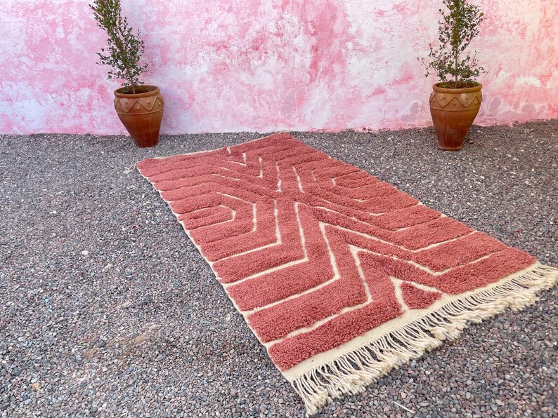 May include: A pink and cream patterned rug with a geometric design. The rug has a fringe border and is laid out on a gravel surface.