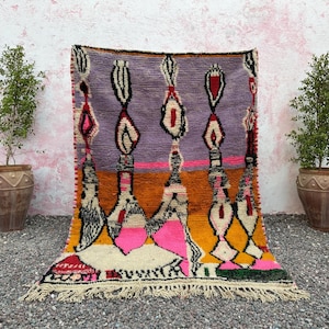 May include: A colorful, hand-woven rug with a geometric design. The rug features a lavender background with vertical patterns in black, white, and red. The lower section has orange, pink, and white geometric shapes. The rug has a fringed edge.