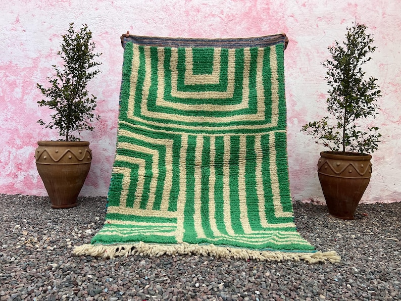 Pode incluir: Um tapete com padr&atilde;o geom&eacute;trico verde e bege com borda com franjas. O tapete &eacute; exibido contra uma parede rosa, ladeado por duas plantas em vasos. O design do tapete apresenta listras verticais e padr&otilde;es quadrados.