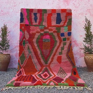 May include: A vibrant, hand-woven rug with a geometric design. The rug features a palette of pinks, reds, oranges, and greens. The rug has a fringed edge and is displayed against a pink wall with potted plants on either side.
