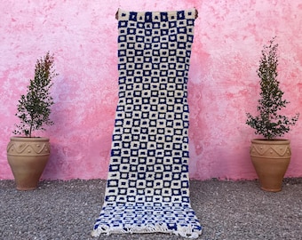 May include: A white and blue geometric patterned rug with a fringe border. The rug is hanging against a pink wall.