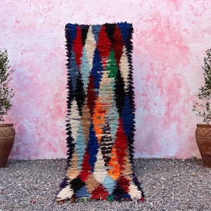 Puede incluir: Una colorida alfombra rectangular con un patrón geométrico en tonos rojos, azules, naranjas y blancos. La alfombra se exhibe contra una pared rosa, con dos plantas en macetas a cada lado. Las dimensiones de la alfombra no son visibles.
