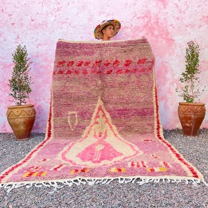May include: A large, rectangular, hand-woven rug with a pink and cream color scheme. The rug features geometric patterns and a central design. The rug has a fringe along the bottom edge and is displayed against a pink wall.
