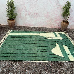 May include: A rectangular green rug with a cream-colored geometric design and black stripes. The rug has a fringe on the edges and is placed on a gravel surface. Two potted plants flank the rug.