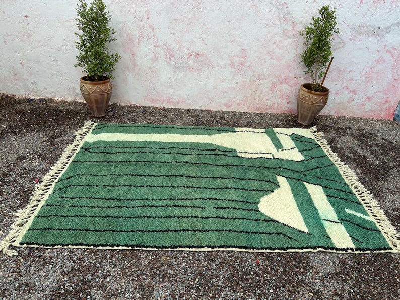 May include: A rectangular green rug with a cream-colored geometric design and black stripes. The rug has a fringe on the edges and is placed on a gravel surface. Two potted plants flank the rug.