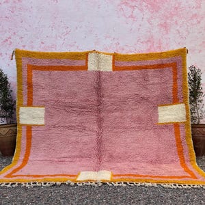 May include: A rectangular, hand-woven rug with a pink central field, bordered by a golden-yellow frame with orange accents. The rug features off-white rectangular details and a fringed edge. The rug is displayed against a pink wall.