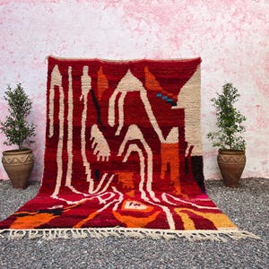 May include: A vibrant, hand-woven rug with a bold geometric design. The rug features a rich red background with cream, orange, and black abstract shapes. The rug is displayed against a pink wall, with potted plants on either side.