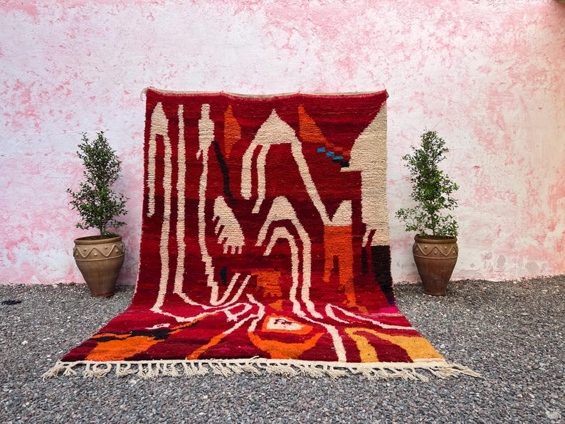 May include: A vibrant, hand-woven rug with a bold geometric design. The rug features a rich red background with cream, orange, and black abstract shapes. The rug is displayed against a pink wall, with potted plants on either side.