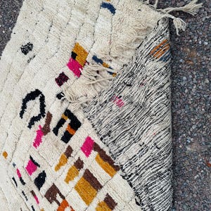 May include: Cream-colored, hand-woven rug with a geometric pattern. The rug features colorful accents in pink, orange, black, and blue. The rug has a textured surface and fringe detailing on the edge. The rug is partially rolled up, revealing a black and white striped pattern.