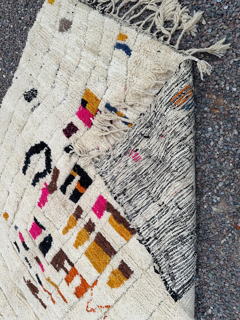 May include: Cream-colored, hand-woven rug with a geometric pattern. The rug features colorful accents in pink, orange, black, and blue. The rug has a textured surface and fringe detailing on the edge. The rug is partially rolled up, revealing a black and white striped pattern.