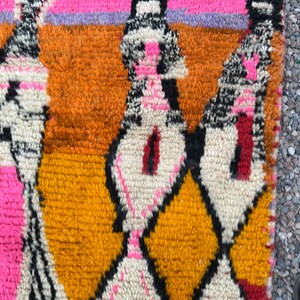 May include: Close-up of a colorful rug with a geometric pattern. The rug features a combination of bright colors, including orange, pink, yellow, and black. The design includes diamond and abstract shapes. The rug is made of a textured material.