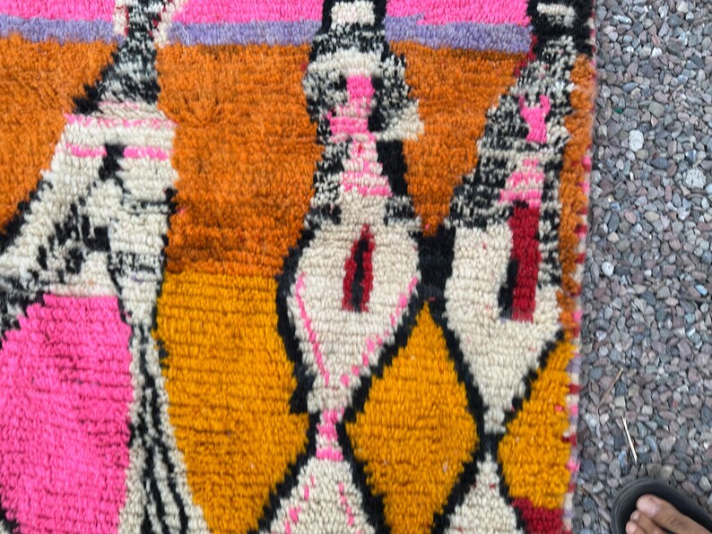 May include: Close-up of a colorful rug with a geometric pattern. The rug features a combination of bright colors, including orange, pink, yellow, and black. The design includes diamond and abstract shapes. The rug is made of a textured material.