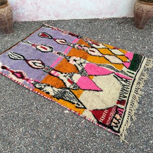 May include: A rectangular, hand-woven rug with a vibrant geometric design. The rug features a palette of purple, orange, yellow, pink, and white, with black and red accents. The rug has a fringed edge and is suitable for home decor.
