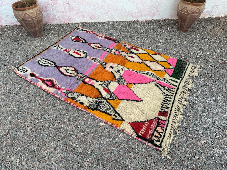 May include: A rectangular, hand-woven rug with a vibrant geometric design. The rug features a palette of purple, orange, yellow, pink, and white, with black and red accents. The rug has a fringed edge and is suitable for home decor.