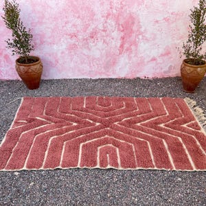 May include: A pink and cream geometric patterned rug with fringe on the edges. The rug is laid out on a gravel surface in front of a pink wall.