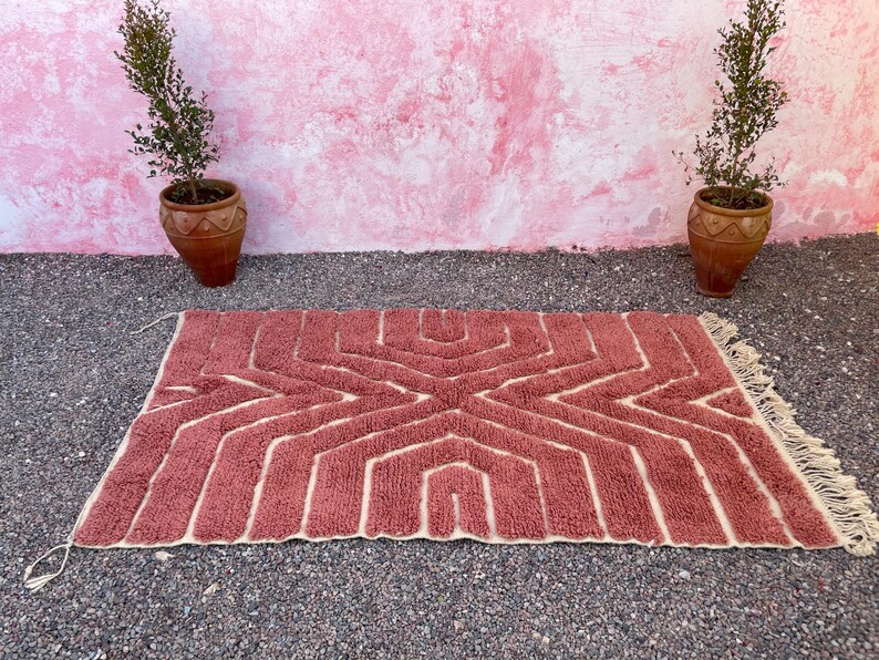 May include: A pink and cream geometric patterned rug with fringe on the edges. The rug is laid out on a gravel surface in front of a pink wall.