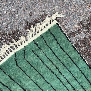 May include: Close-up of a green rug with black vertical stripes. The rug has a cream-colored border with fringe. The rug is made of a soft, textured material. The rug is placed on a surface of small, gray stones.