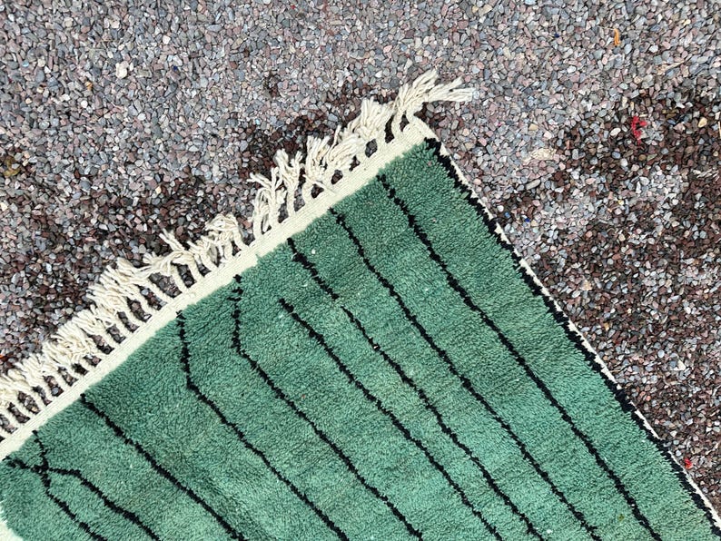 May include: Close-up of a green rug with black vertical stripes. The rug has a cream-colored border with fringe. The rug is made of a soft, textured material. The rug is placed on a surface of small, gray stones.