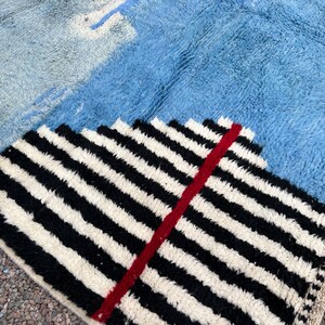 May include: A geometric rug with a textured, shaggy pile. The rug features sections of light blue, medium blue, and a black and white striped pattern with a red accent. The rug has a fringed edge.