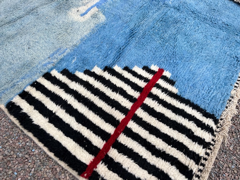 May include: A geometric rug with a textured, shaggy pile. The rug features sections of light blue, medium blue, and a black and white striped pattern with a red accent. The rug has a fringed edge.
