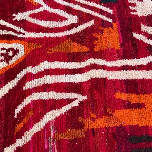 May include: Close-up of a vibrant, textured rug with a bold geometric pattern. The rug features a rich red background with white, orange, and yellow accents. The design includes wavy lines and angular shapes, creating a striking visual effect.