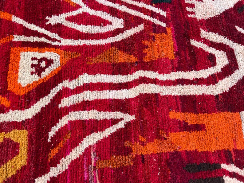 May include: Close-up of a vibrant, textured rug with a bold geometric pattern. The rug features a rich red background with white, orange, and yellow accents. The design includes wavy lines and angular shapes, creating a striking visual effect.