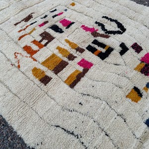 May include: Cream-colored, textured rug with a geometric pattern. The design features blocks of color in shades of brown, orange, yellow, pink, and black. The rug has a rectangular shape and a thick pile.