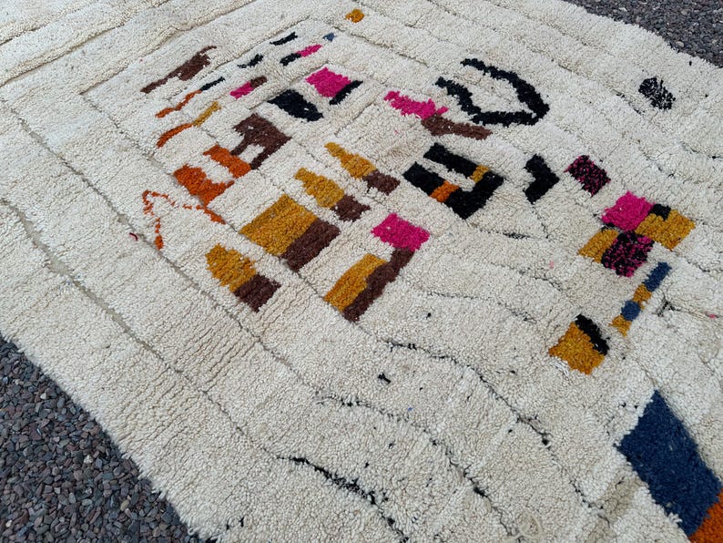 May include: Cream-colored, textured rug with a geometric pattern. The design features blocks of color in shades of brown, orange, yellow, pink, and black. The rug has a rectangular shape and a thick pile.