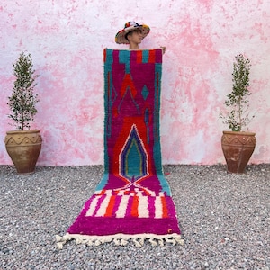 May include: A long, colourful Moroccan rug with a geometric pattern in shades of pink, red, blue, and white. The rug is being held up by a person wearing a hat.