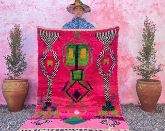 May include: A pink and green Moroccan rug with geometric patterns. The rug is being held up by a person wearing a colorful hat.