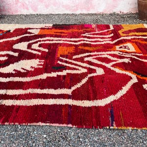 May include: A rectangular, hand-woven rug with a vibrant abstract design. The rug features a rich red base with white, orange, and black accents. The design includes flowing white lines and geometric shapes, creating a bold and artistic statement. The rug is made of a thick, textured material.