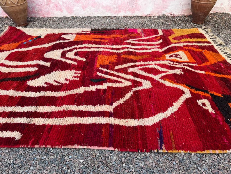 May include: A rectangular, hand-woven rug with a vibrant abstract design. The rug features a rich red base with white, orange, and black accents. The design includes flowing white lines and geometric shapes, creating a bold and artistic statement. The rug is made of a thick, textured material.