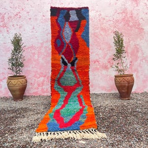Pode incluir: Um tapete abstrato colorido com um padrão geométrico em tons de vermelho, laranja, azul, verde e branco. O tapete tem uma borda de franjas e é exibido contra uma parede rosa.
