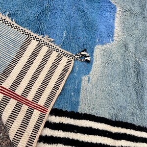 May include: A blue and white rug with a geometric design. The rug features a combination of light and dark blue hues, with sections of white and black stripes. The edge of the rug has a striped pattern with tassels.