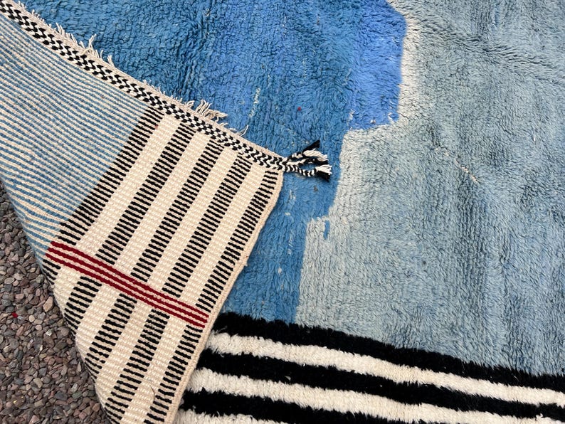 May include: A blue and white rug with a geometric design. The rug features a combination of light and dark blue hues, with sections of white and black stripes. The edge of the rug has a striped pattern with tassels.