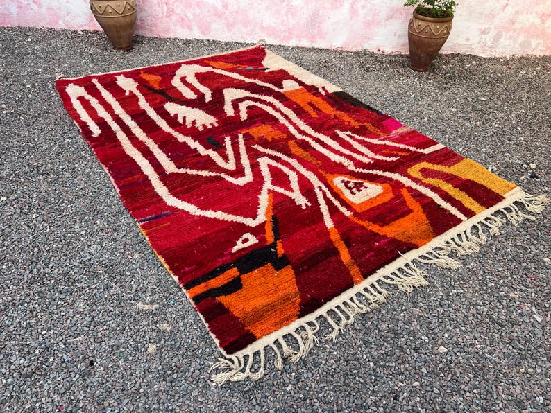May include: A rectangular, hand-woven rug with a vibrant, abstract design. The rug features a rich palette of red, orange, and white, with fringe along the edges. The design includes geometric shapes and lines.