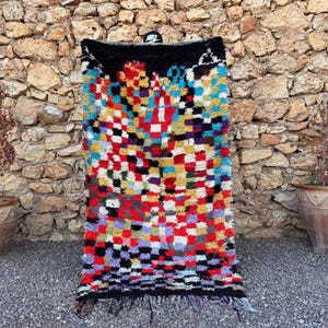 May include: A rectangular wool rug featuring a vibrant, multi-colored checkered pattern in shades of red, blue, yellow, green, and purple. The rug has a solid black border at the top and bottom, with a fringed edge at the base.
