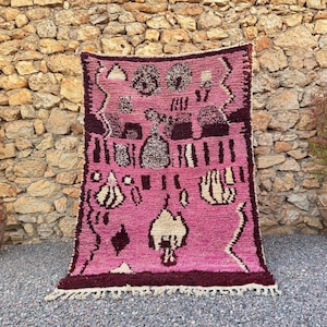 May include: A rectangular wool rug featuring a vibrant pink background with dark purple and cream-colored geometric patterns and abstract shapes. The rug has a thick, shaggy texture and includes a fringed edge at the bottom with knotted details.