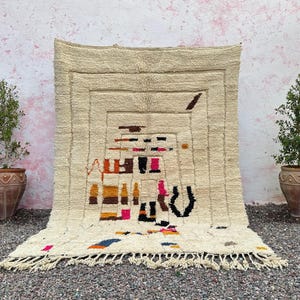 May include: Cream-colored, rectangular rug with a geometric design in shades of brown, black, pink, and orange. The rug has a fringed edge and is displayed against a textured wall. Two potted plants flank the rug.