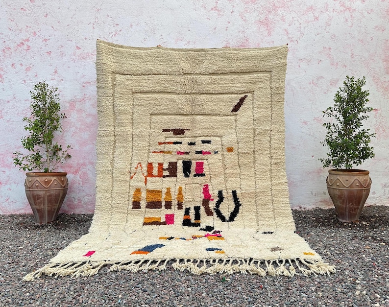 May include: Cream-colored, rectangular rug with a geometric design in shades of brown, black, pink, and orange. The rug has a fringed edge and is displayed against a textured wall. Two potted plants flank the rug.